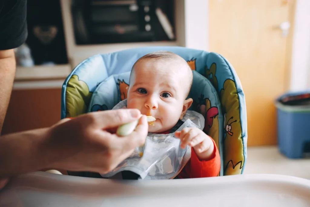 Bébé assis dans une chaise haute avec une cuillère dans la bouche donnée par un adulte qu'on ne voit pas 